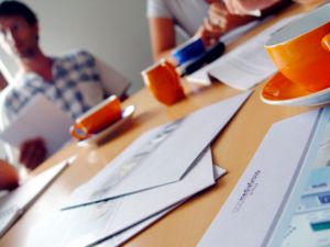 Ein runder Tisch in einer Agentur. Rundherum sitzen Mitarbeiter bei einem Meeting. Auf dem Tisch stehen orangene Tassen und liegen firmeninterne Unterlagen. Wie bei der Web-Usability, trotz der Menge an Objekten wird die Übersichtlichkeit gewahrt.